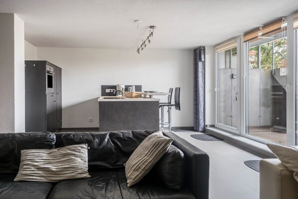 A modern living room with a black leather sofa, a kitchen area with a dining table, and a large sliding glass door leading to an outdoor space.