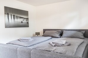 A neatly made bed with grey linens and pillows, a small bedside table with a lamp, and a large abstract painting on the wall.