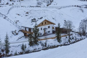 A snow-covered house with a wooden exterior and a name plaque, surrounded by snow-covered trees and a stone wall.