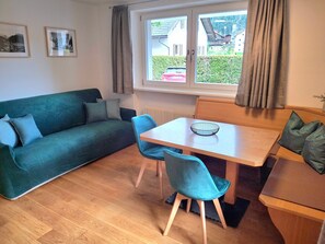 Une pièce avec un canapé vert, deux fauteuils bleus, une table en bois sur laquelle se trouve un saladier, une fenêtre avec des rideaux et des tableaux encadrés accrochés au mur.