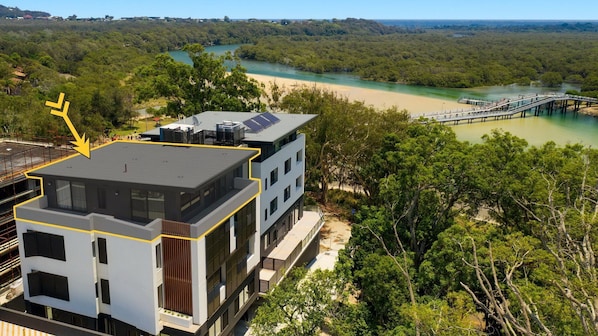 A modern multi-story building with a flat roof, surrounded by dense greenery and a river.