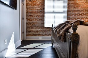 A room with a brick wall, a bed with a wooden headboard, and a window with blinds.