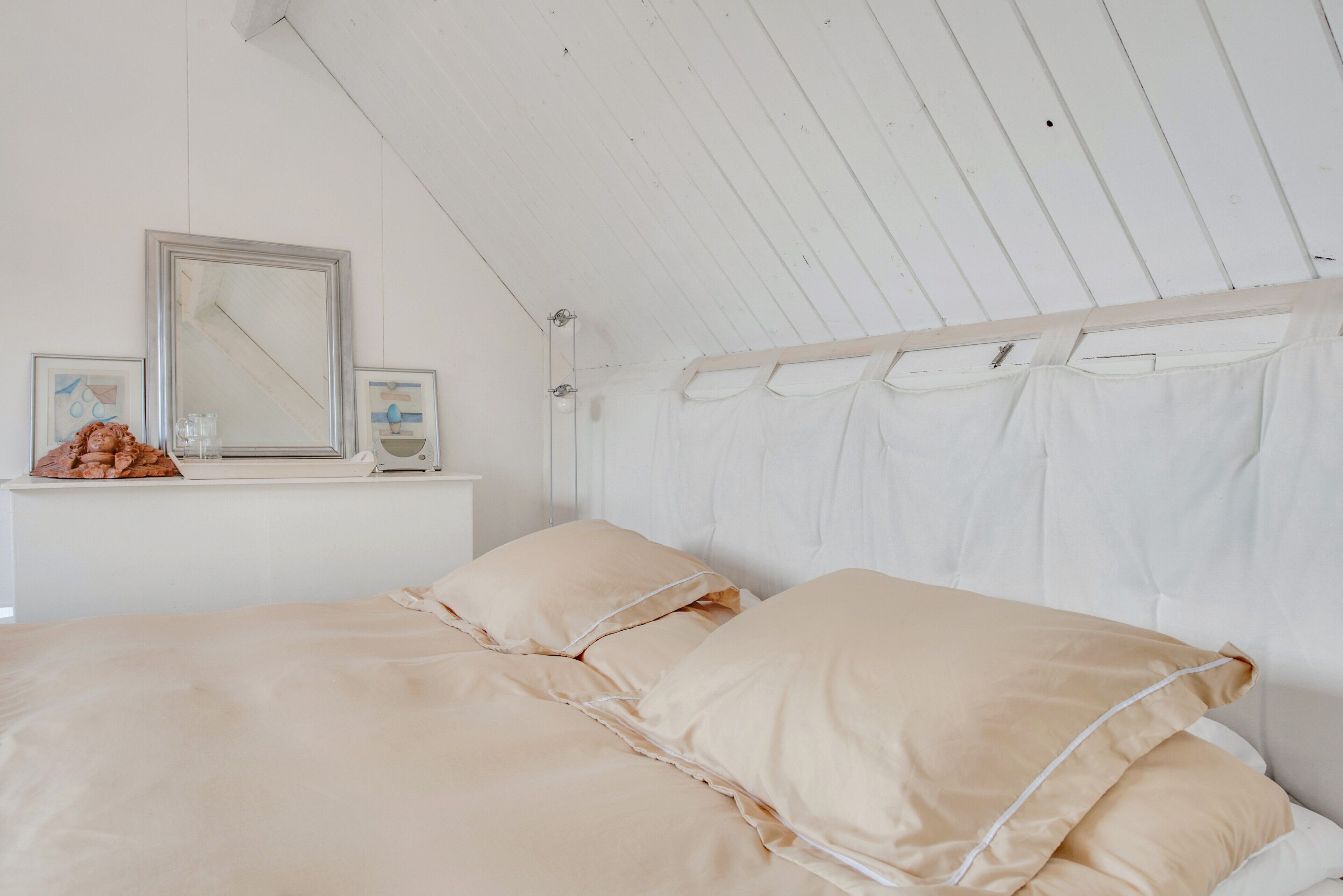A bedroom with a sloped ceiling, a bed with beige bedding, a bedside table with a mirror and framed pictures, and a window with white curtains.