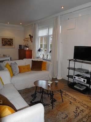 A modern living room with a sofa, a coffee table, a TV stand with electronic devices, and a window with curtains. - Holiday Home "Engel van de Molen" for Couples with Terrace, Garden & Wi-Fi (HOOGWOUD)
