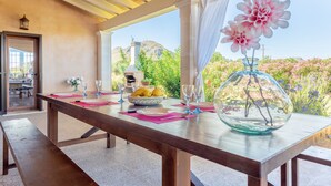 A dining table set with plates, cutlery, and a bowl of fruit, with a view of a mountain and flowers. - Finca "S'Hortet" with Sea View, Private Pool & Wi-Fi (Alcúdia)