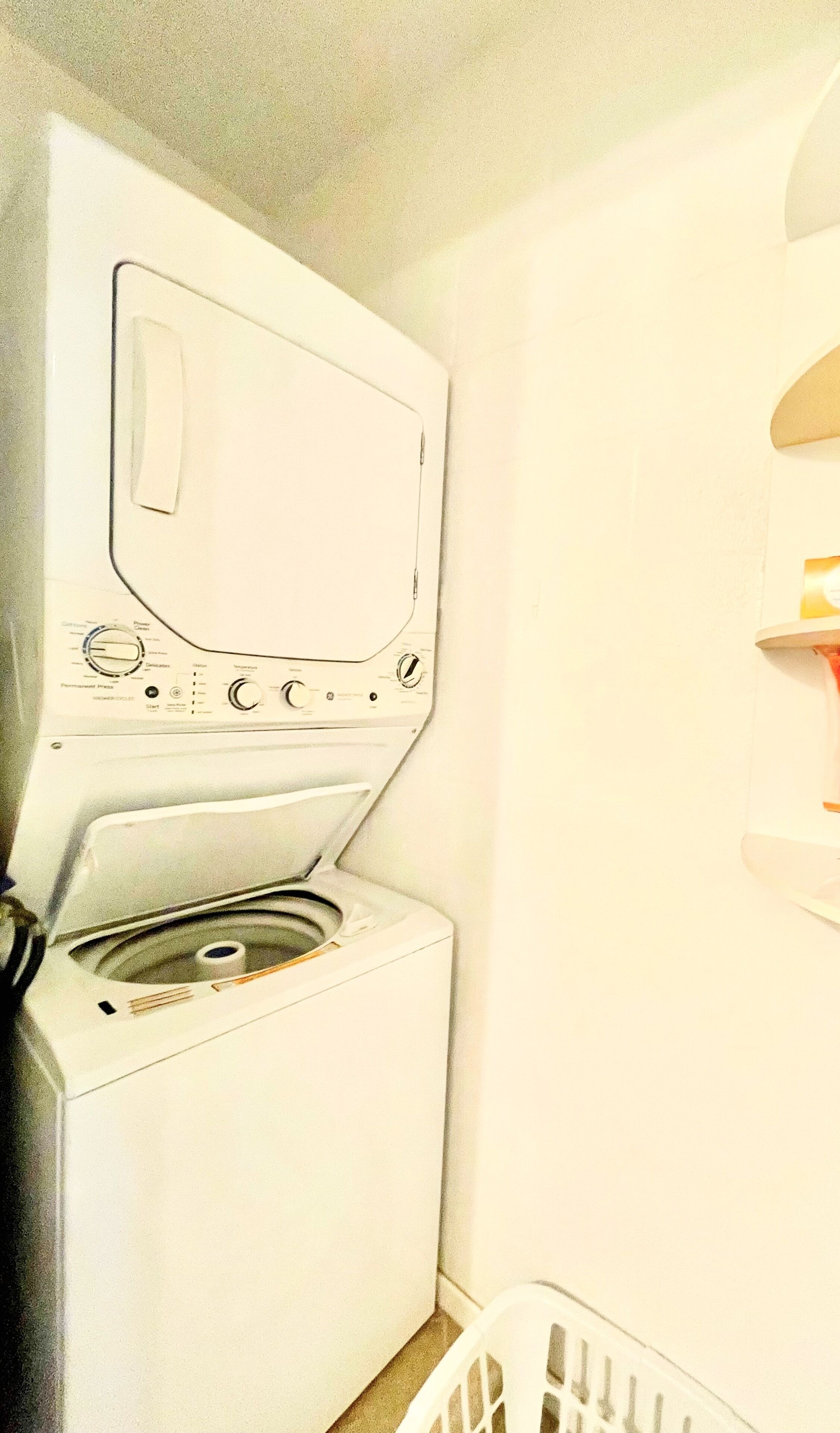 A front-loading washing machine with a control panel, a basket, and a wall-mounted shelf.