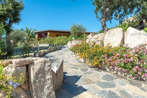 A stone pathway leading to a building with a tiled roof, surrounded by large rocks and flowering plants. - Residence Villa Smeralda 3 (Telti)