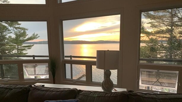 A room with a view of a sunset over water, a sofa with a cushion, and a lamp on a table. - Luxury Cottage on ice-fishing Rice Lake and year round activities (Bewdley)