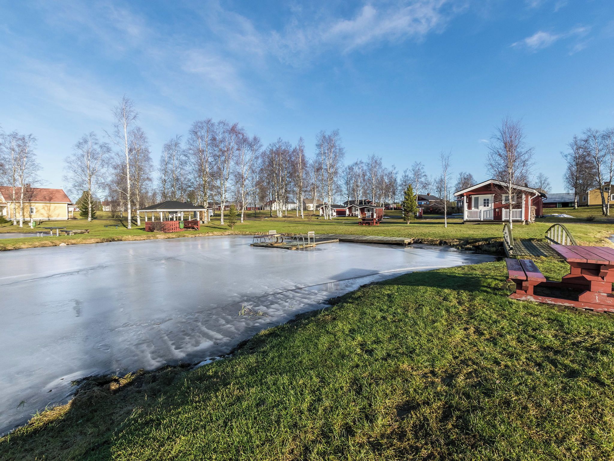 A frozen pond with a gazebo, picnic tables, and houses in the background.
