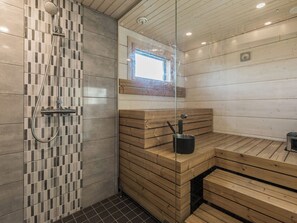 Un sauna con bancos de madera y puerta de vidrio, zona de ducha con paredes de azulejos y una pequeña ventana sobre los bancos.
