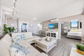 Uma sala de estar moderna com sofá, mesa de centro e TV. Há uma área de jantar com mesa e cadeiras, e um quarto com cama e janela.