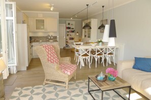 A cozy living room with a wicker chair, a dining table with chairs, a sofa with a blue pillow, and a coffee table with decorative items.