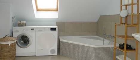 A laundry area with a washing machine and dryer, a corner bathtub, and a wooden shelf with jars.