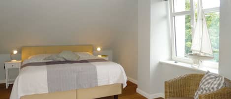 A bedroom with a bed, a nightstand, a chair, a window with curtains, and a view of trees outside.
