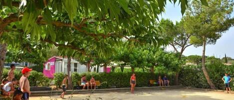 A sandy outdoor area with people sitting and standing, surrounded by greenery and a pink building.