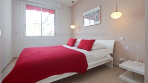 A modern bedroom with a bed, red pillows, a white headboard, a window with a red curtain, and a wall-mounted artwork. - Montrachet-2429-Modern 2 bed rooms in the city center (Cannes)