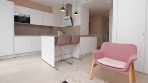 A modern kitchen with white cabinets, a built-in oven, and a dining area with a pink chair. - Montrachet-2429-Modern 2 bed rooms in the city center (Cannes)