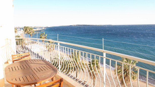 Een balkon met een houten tafel en stoelen, uitkijkend op een strand met palmbomen en de zee.