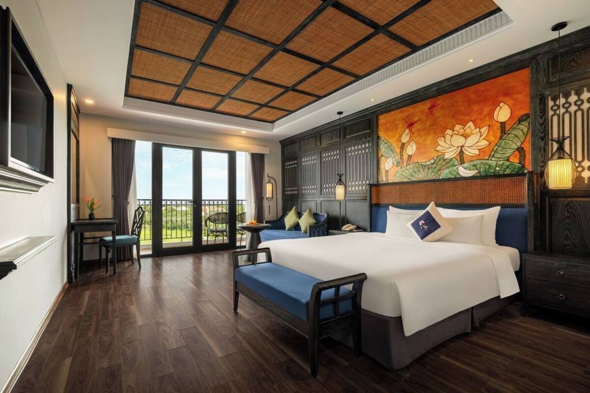 A hotel room with a large bed, a desk with a chair, a balcony with a view, and a decorative wall with a lotus painting.