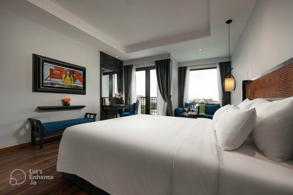 A hotel room with a large bed, a desk with a chair, a sofa, a TV mounted on the wall, and a window with curtains.