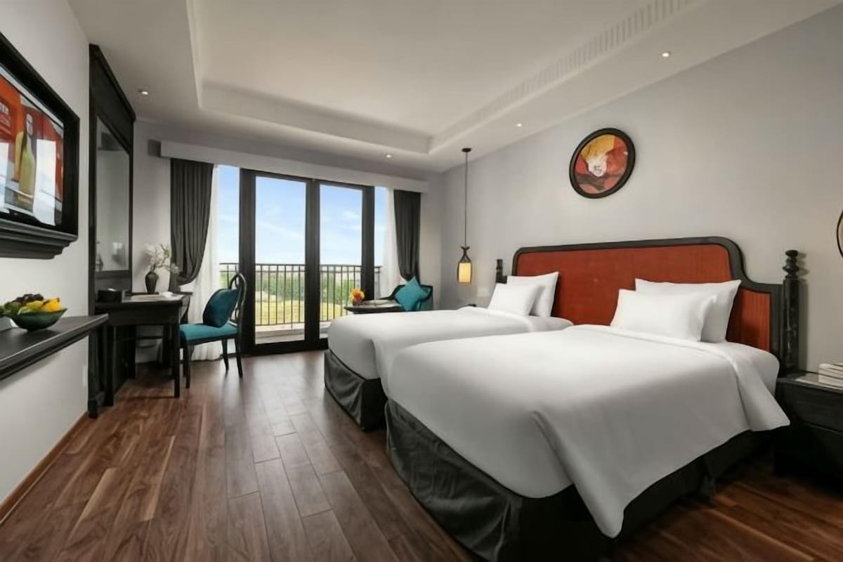 A hotel room with two beds, a desk with a chair, a balcony, and a large window.