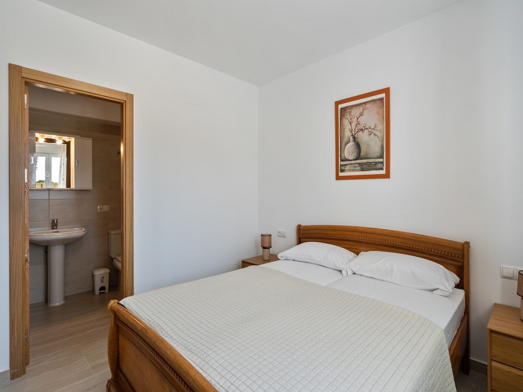 A bedroom with a wooden bed, a bedside table, a nightstand, a wall-mounted lamp, and a framed picture.