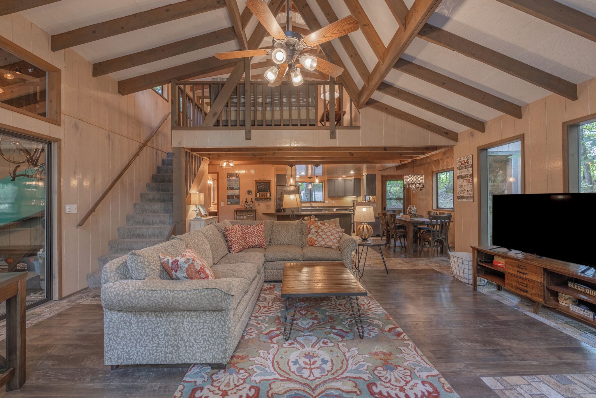 A spacious living room with a large sectional sofa, a wooden coffee table, and a patterned rug. A staircase leads to an upper level with a balcony.