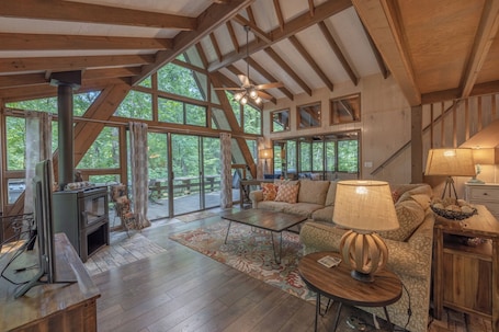 A spacious living room with a large fireplace, wooden beams, and large windows offering a view of the surrounding forest.
