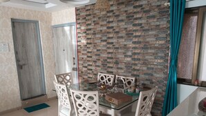 A dining area with a glass table and white chairs, a brick-patterned wall, a wooden door, and a teal curtain.