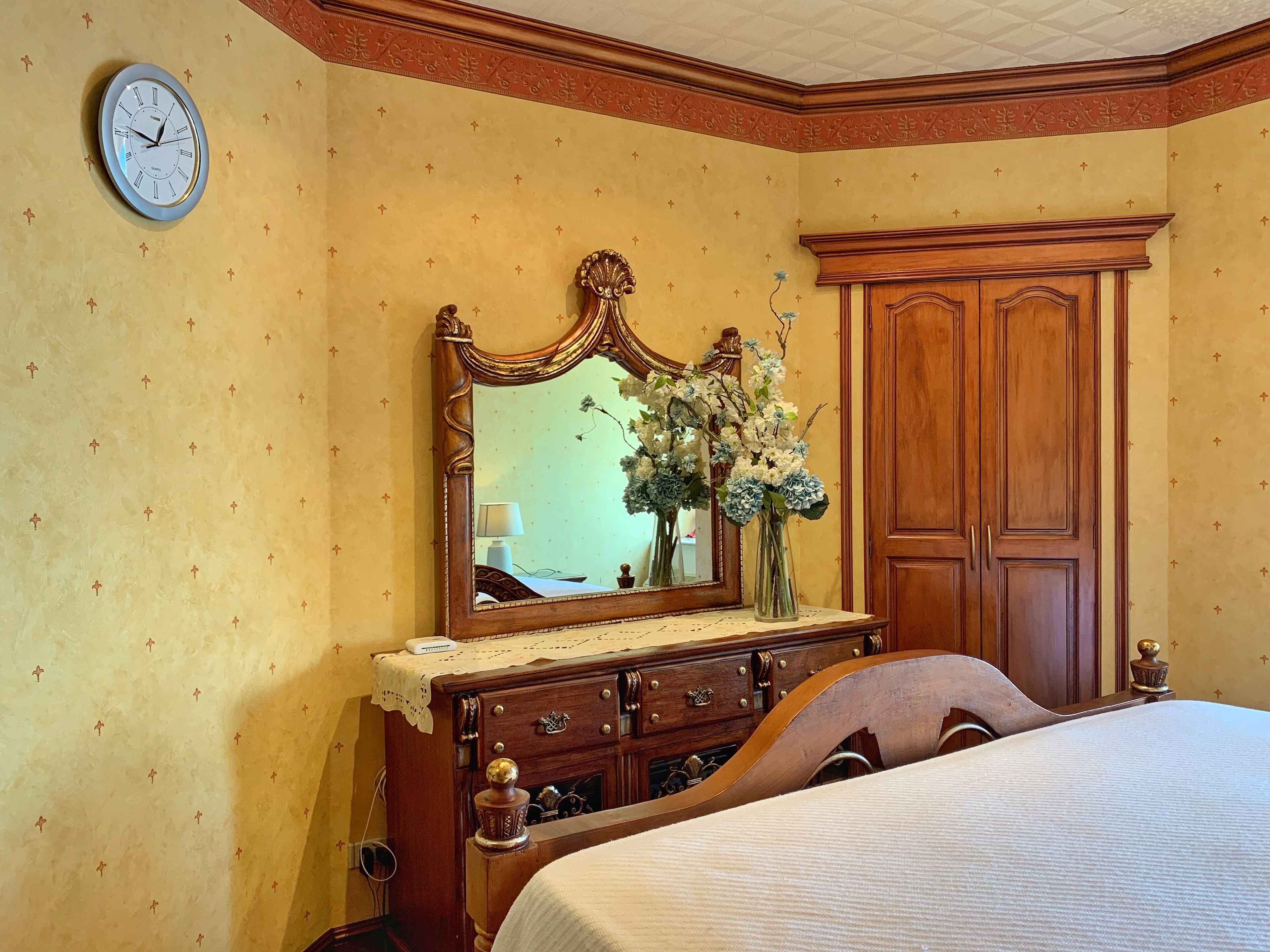 Ein Schlafzimmer mit einem hölzernen Bett, einer Kommode mit Schubladen, einem Spiegel mit Blumenarrangement und einer Wanduhr.