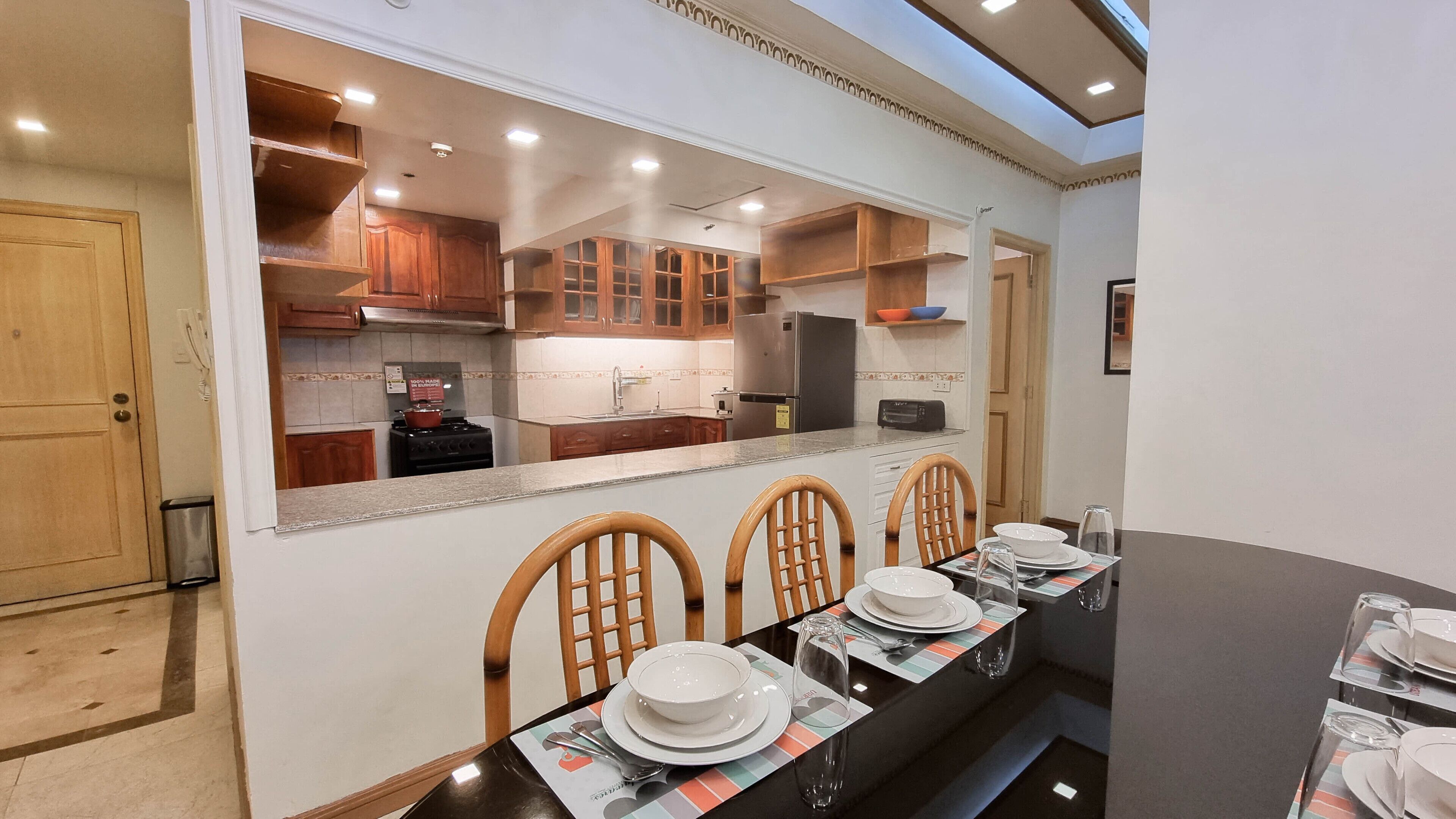 A dining area with a long table set for four, wooden chairs, and a kitchen visible through a glass partition.