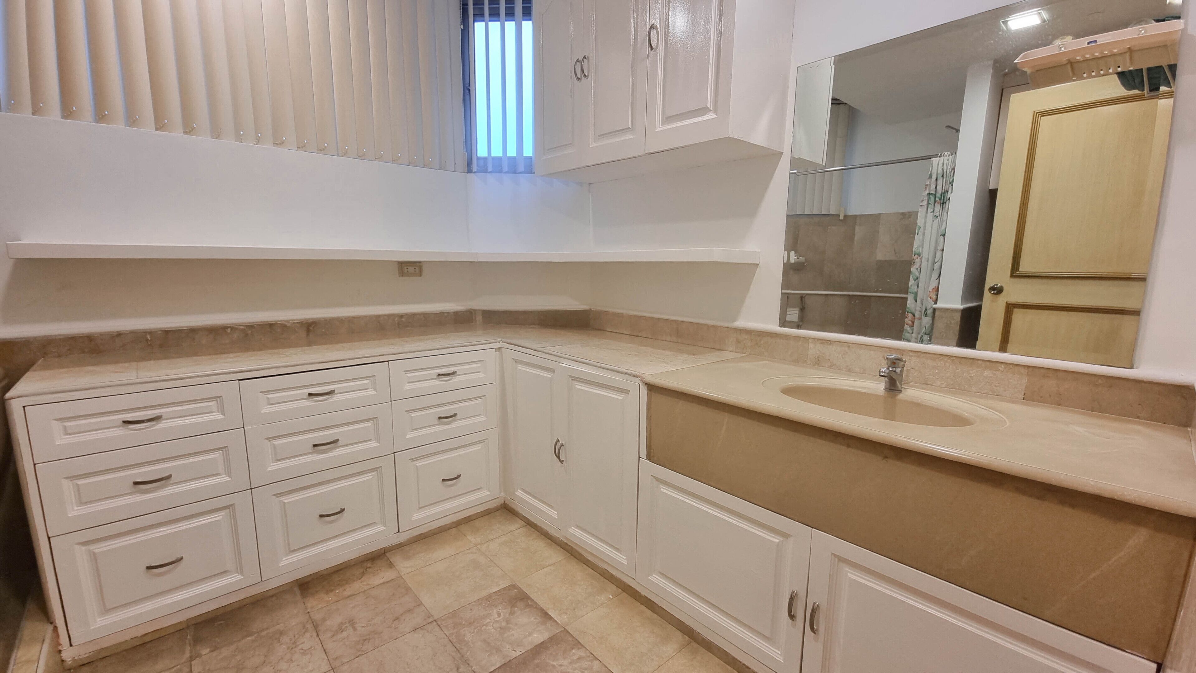 A bathroom with a beige tiled floor, white cabinetry, a sink with a mirror above it, and a shower visible in the reflection.