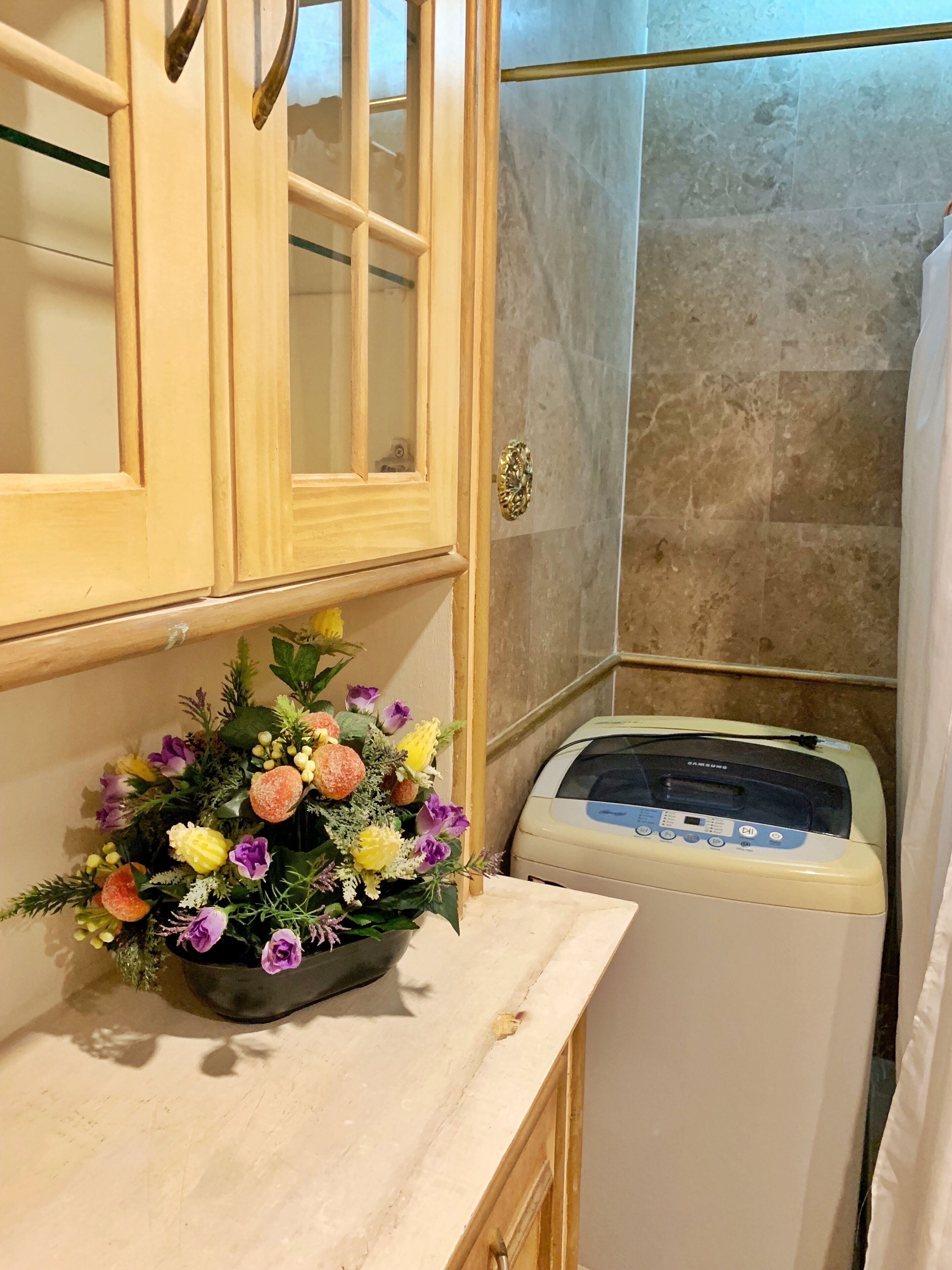 A bathroom with a wooden cabinet, a wall-mounted washing machine, and a floral arrangement on the counter.