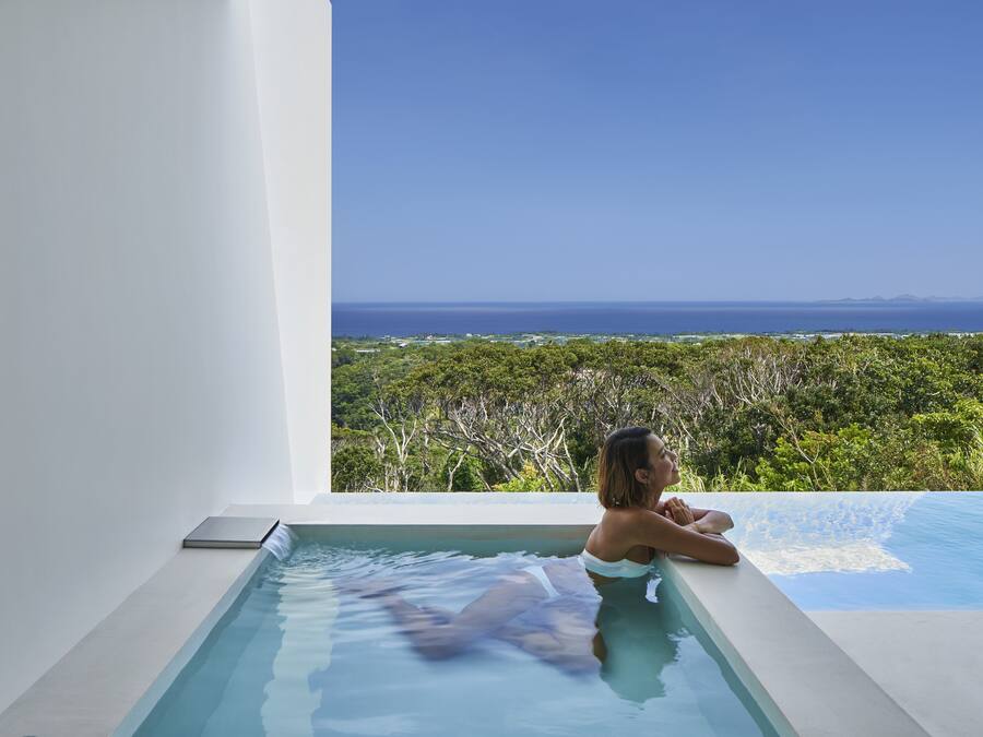 A woman relaxing in a modern infinity pool with a view of the ocean and greenery.