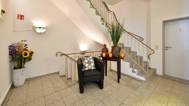 A hotel lobby with a staircase, a black armchair with a patterned cushion, a potted plant, and a small table with candles.