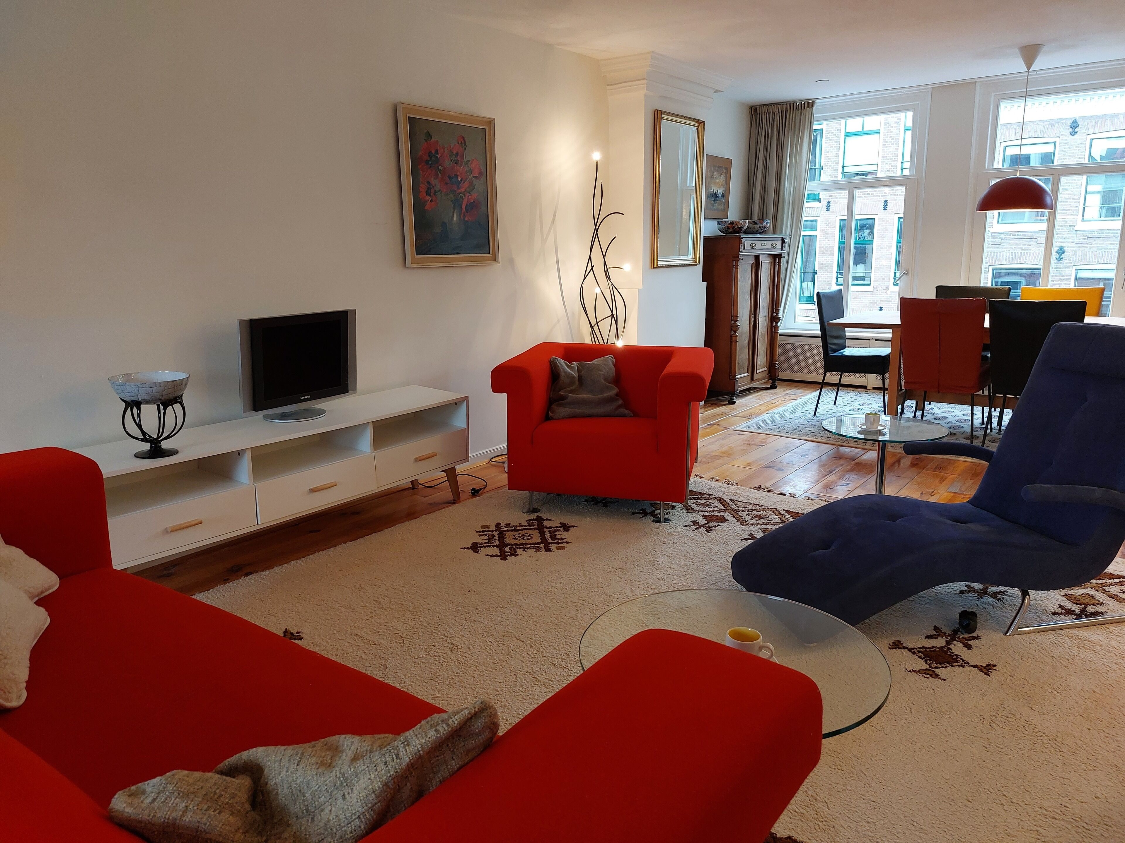 A living room with a red sofa, a blue armchair, a glass coffee table, and a TV on a white stand.