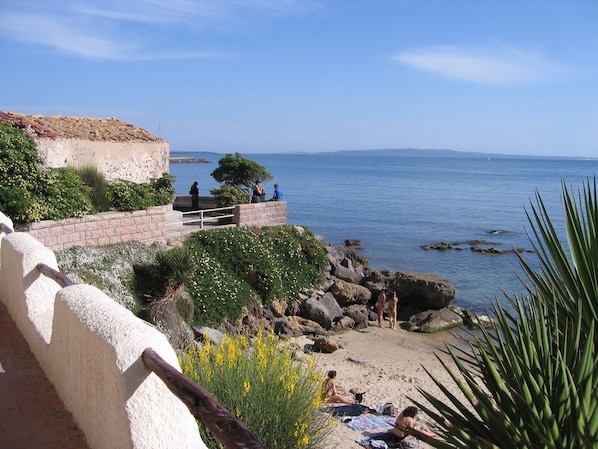 A coastal walkway with a stone wall, a small building, and a beach with people and vegetation.