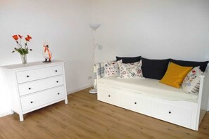 Un canapé blanc agrémenté de coussins à motifs floraux et d’un coussin jaune, une commode blanche avec des tiroirs et un lampadaire.