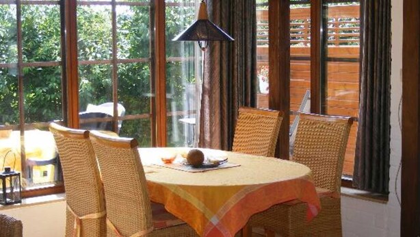 Een eetgedeelte met rieten stoelen en een ronde tafel, grote ramen met gordijnen en een hanglamp.