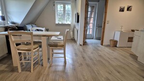 Una habitación con una mesa de comedor, dos sillas, un radiador, un televisor montado en la pared y una ventana con cortinas.