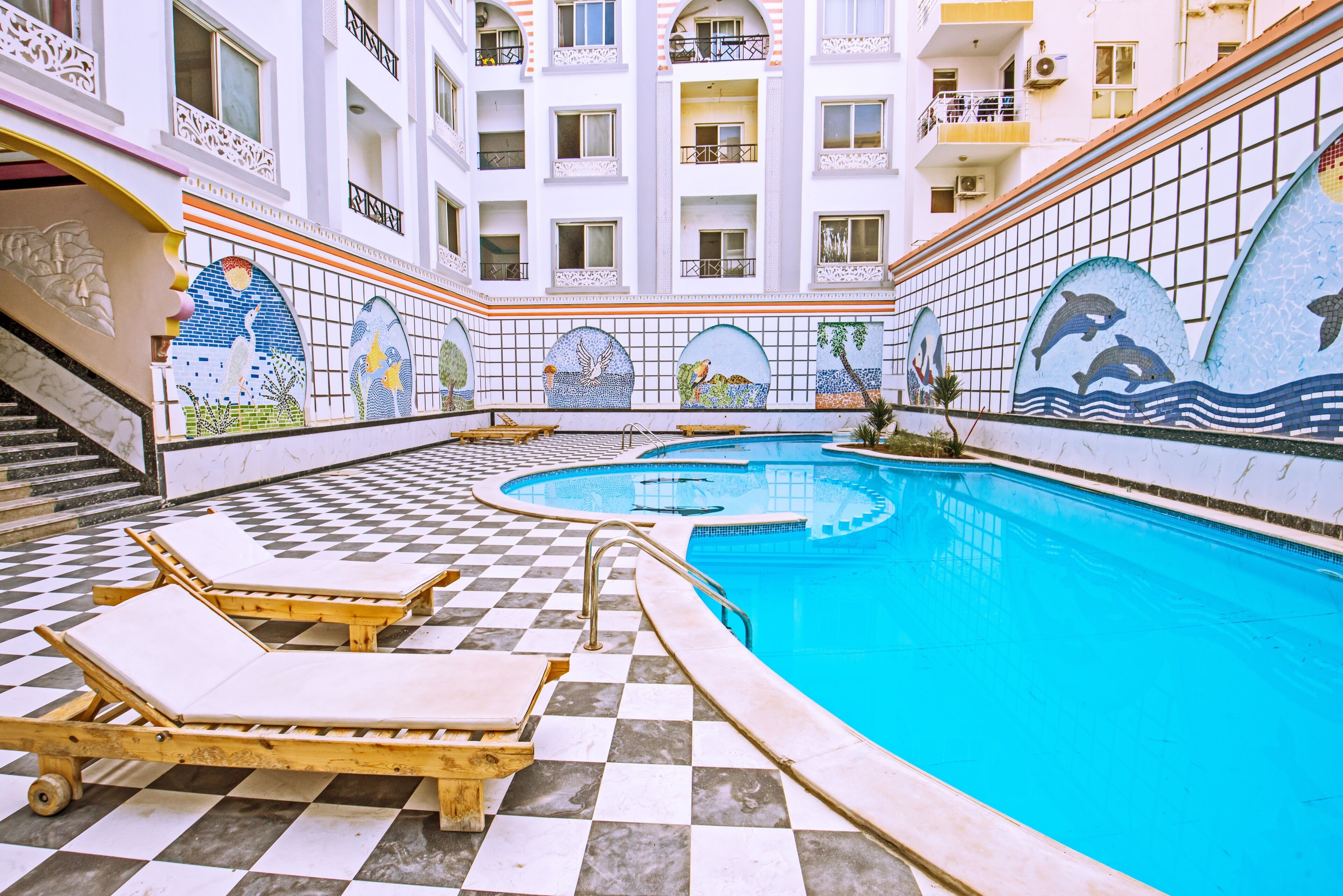 Ein Schwimmbecken mit Schachbrettboden und Liegestühlen, umgeben von einem Gebäude mit Balkonen und dekorativen Mosaiken.