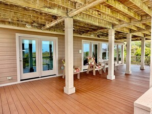 A covered outdoor deck with wooden flooring, white pillars, and glass doors leading to a balcony.
