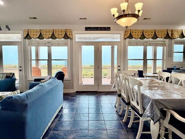 A dining area with a view of the beach, a sign reading 'Come Gather At Our Table', and a chandelier.