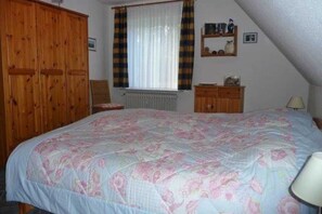 Un dormitorio con una cama grande, un armario de madera, un radiador, una ventana con cortinas y un estante con objetos.