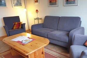 Un salón con un sofá gris, una mesa de centro de madera con papeles, un sillón azul con un cojín a rayas y una lámpara de pie.
