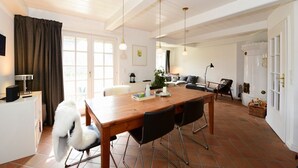 A spacious dining area with a wooden table and chairs, a white ceiling, and a view of the living room in the background.
