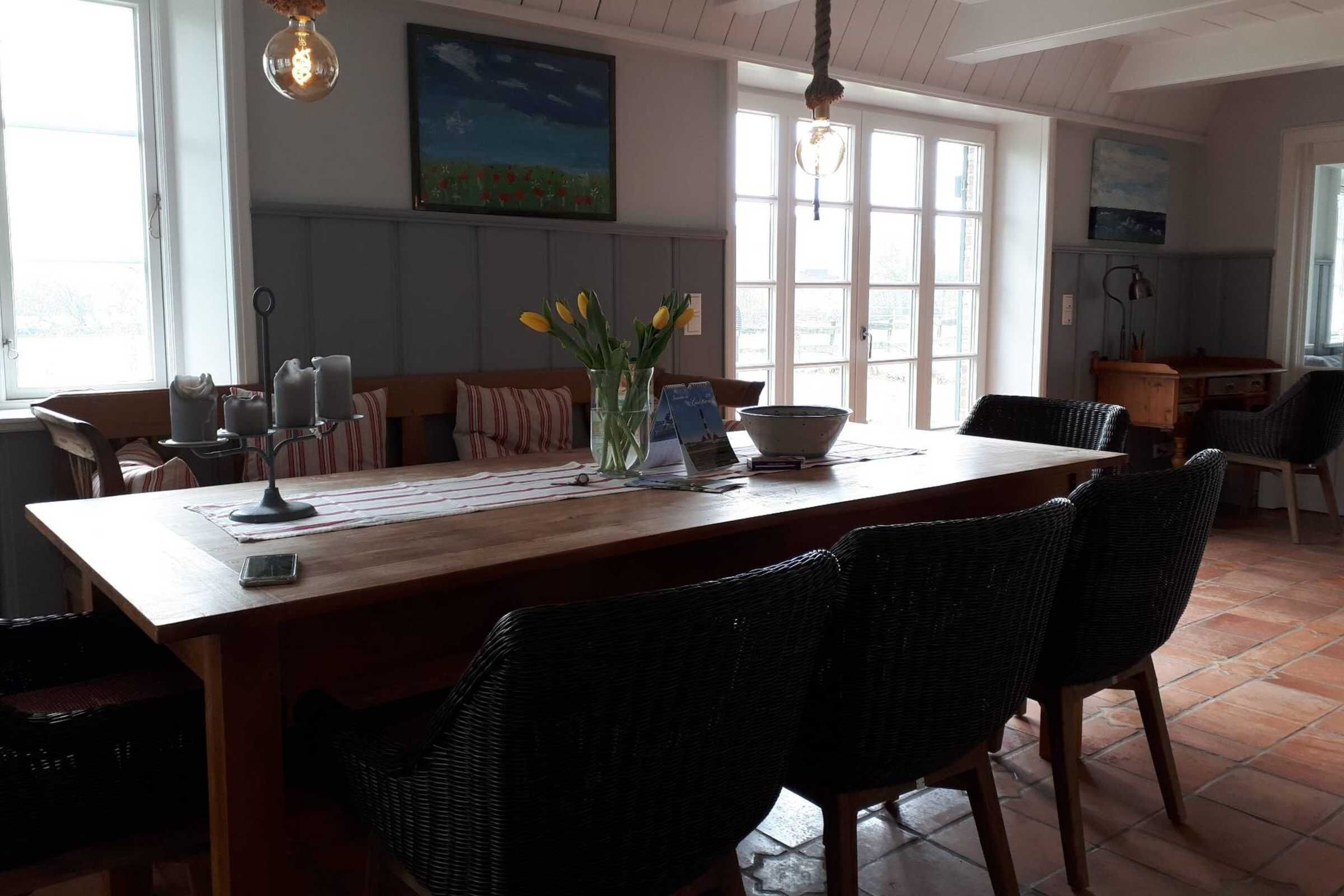 En spiseplads med et træbord, fletstole, en vase med blomster og et billede på væggen.