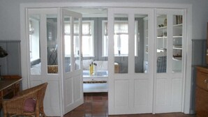 Quarto com porta branca que dá para um quarto, uma cadeira de vime, piso de azulejos e uma estante.