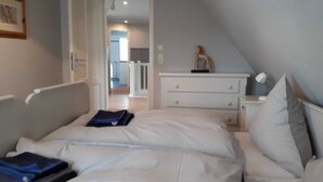 Quarto com cama, mesa de cabeceira, cômoda e porta que dá para outro espaço.