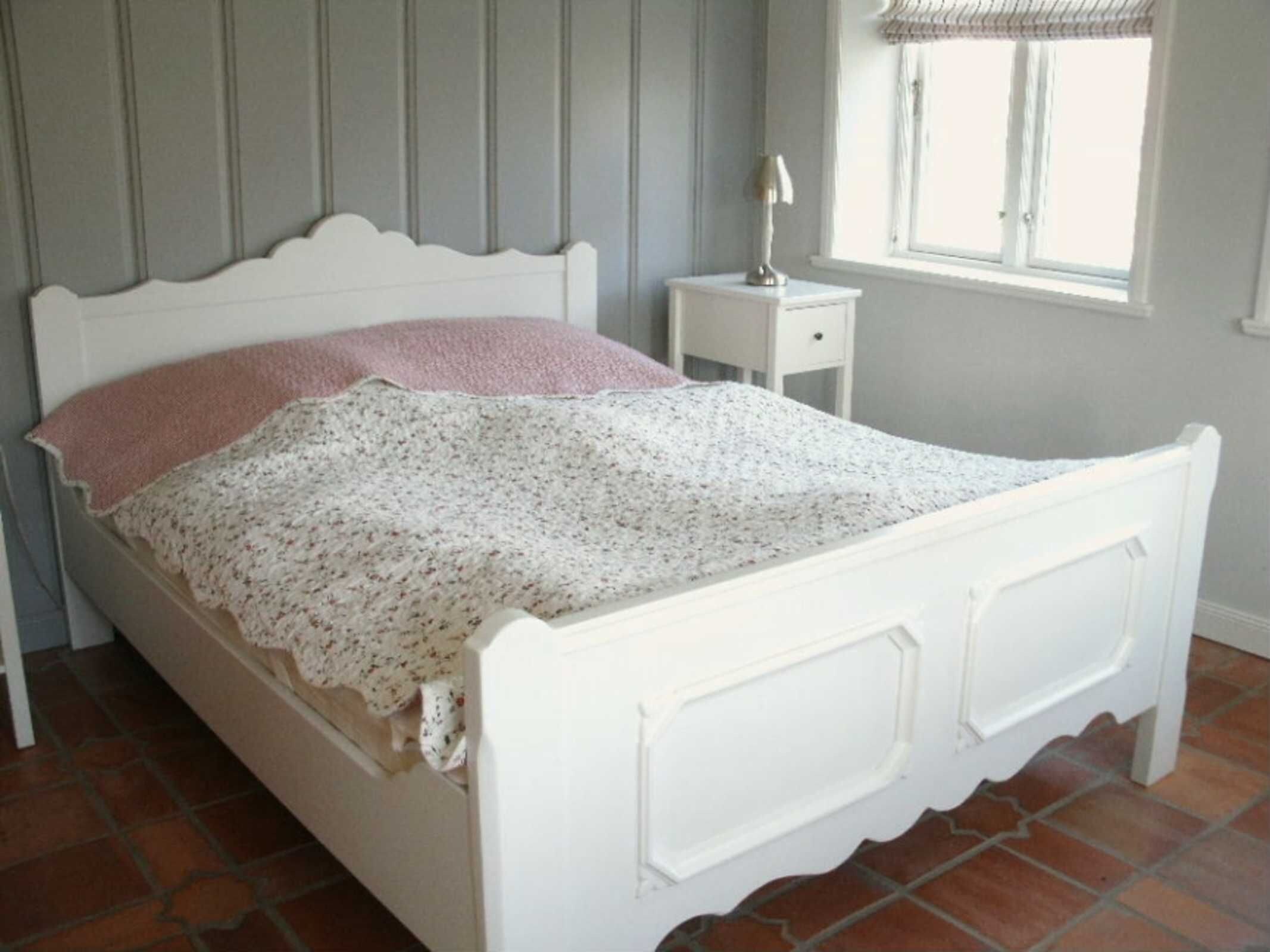 Un lit blanc avec une couvre-lit à motifs floraux et un oreiller rose, dans une pièce aux murs gris et dotée d’une fenêtre avec des stores.